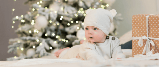 baby in white woollen hat in front of the christmas tree